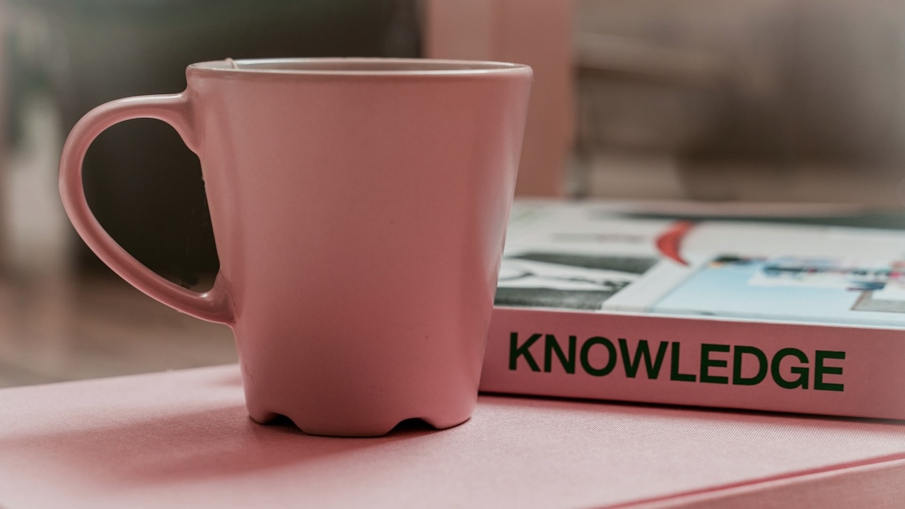 Pink mug on a table next to a book with the word knowledge written on the spine