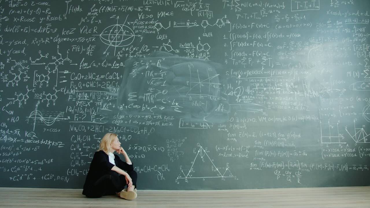Lady sitting on the floor in front of a large chalk board that is displaying a many calclations